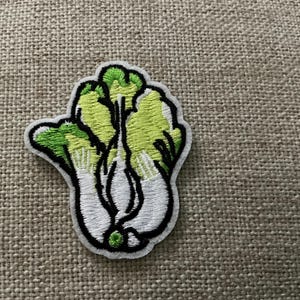 May include: An embroidered patch shaped like a head of bok choy. The vegetable features white stems at the base transitioning into light green and dark green leafy tops, all outlined with a thick black border on a white fabric backing.