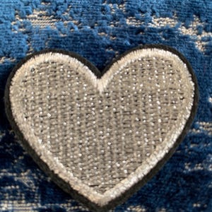 May include: A silver heart-shaped patch with a black border. The patch is made of a sparkly material and has a slightly raised texture.