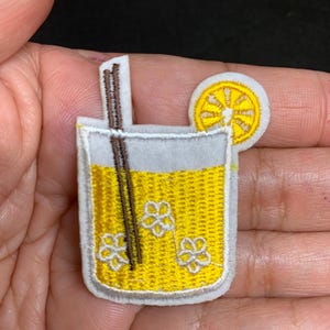 May include: Embroidered patch featuring a yellow drink in a glass with a lemon slice and a straw. The drink has white floral accents and a gray rim. The patch is held in a hand.