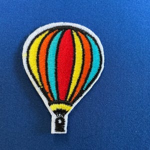 May include: Embroidered hot air balloon patch with a white border. The balloon features vertical stripes in yellow, orange, red, and teal. The bottom of the balloon and the basket are black. The patch is on a blue background.