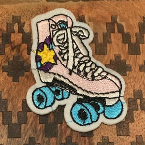 May include: A pink roller skate with blue wheels and a yellow star on the side. The skate is embroidered on a white felt patch.