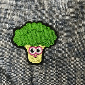 May include: Embroidered patch featuring a cartoon broccoli character with large eyes and a smiling face. The broccoli head is green, the stem is yellow, and the outline is black. The patch is on a blue and white textured fabric.