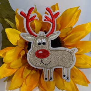 May include: An embroidered patch shaped like a reindeer with a tan body, a bright red nose, and red antlers. The reindeer has white eyes with black pupils and a simple stitched mouth. The patch features a white border outline throughout.