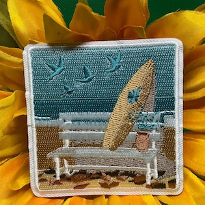 May include: Embroidered patch featuring a beach scene with a surfboard, bench, and flying birds. The patch has a square shape with a white border. The background is a teal color, and the surfboard is tan.