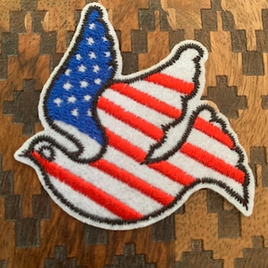 May include: Embroidered patch in the shape of a dove, featuring the American flag design. The dove's wings display the blue field with white stars and red and white stripes. The patch has a black border and is set against a wooden background.