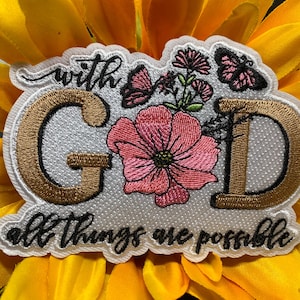 May include: An embroidered patch featuring the text with GOD all things are possible in gold and black thread. The design includes a large pink flower, smaller pink floral accents, and two black butterflies on a white background with a scalloped edge.