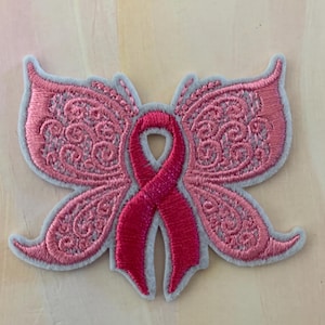 May include: Pink embroidered butterfly with a pink ribbon in the center. The butterfly has a lacy design.