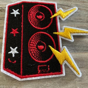 May include: Embroidered patch featuring a black speaker design with red speaker cones and accents, white stars, and three yellow lightning bolts. The patch has a white border and is likely for clothing or accessories.