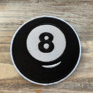 May include: Embroidered patch featuring the number 8 from a pool ball. The patch is circular, with a black outer ring and a white inner circle. The number 8 is black, and a white curved line is below it.