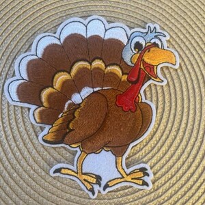 May include: Embroidered patch of a cartoon turkey with brown feathers, a red wattle, and a white head. The turkey is smiling and has large, round eyes.