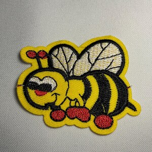 May include: Embroidered patch featuring a cartoon bee with a yellow body, black stripes, and white wings. The bee has a smiling face with red accents and is surrounded by a yellow border. The patch is designed for application on clothing or accessories.