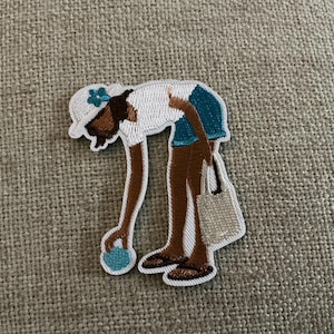 May include: An embroidered patch depicting a figure bending over to pick up a small blue object. The figure wears a white hat with a blue flower, a white top, blue shorts, and brown sandals, while carrying a small white tote bag on one arm.