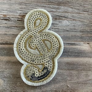 May include: A white felt treble clef patch, embellished with gold and silver sequins. The musical note-shaped patch has a gold embroidered outline and is set against a weathered wood background. A decorative item for music lovers.