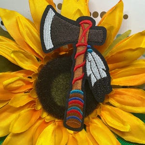May include: An embroidered patch featuring a brown axe with a silver blade, a red wrapped handle, and two white feathers with blue and black accents attached to the side. The handle base has horizontal stripes in blue, red, and brown colors.