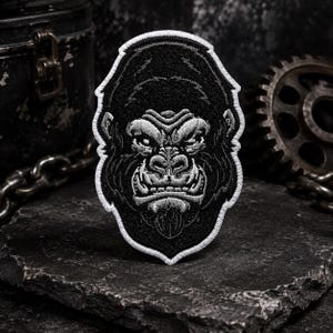 May include: An embroidered patch featuring a detailed black and white illustration of a gorilla face. The design is rendered in high-contrast thread, showcasing the primate's intense expression, textured fur, and facial features, finished with a white border.