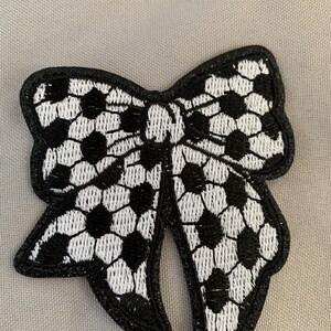 May include: Black and white embroidered bow with a lace-like pattern. The bow is made of fabric and has a black outline.