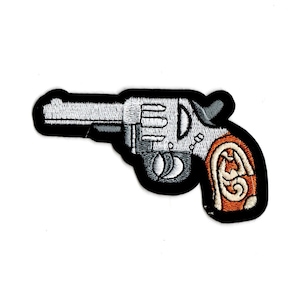 May include: A silver and black embroidered patch of a revolver with a brown and white patterned handle.