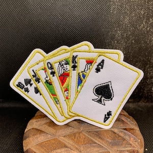 May include: A set of playing card patches featuring the Ace of Spades, Queen, Jack, and King of clubs and diamonds. Each card has a white background with black and red details, outlined in gold. The patches are displayed on a wooden surface.