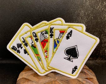 May include: A set of playing card patches featuring the Ace of Spades, Queen, Jack, and King of clubs and diamonds. Each card has a white background with black and red details, outlined in gold. The patches are displayed on a wooden surface.