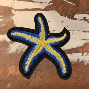 May include: A blue and yellow embroidered starfish patch with a black outline. The patch is perfect for adding a nautical touch to clothing, bags, or other crafts.