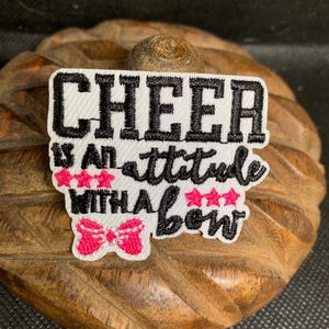 May include: A white patch with black embroidered text that reads "CHEER IS AN ATTITUDE WITH A BOW." The patch features pink stars and a pink bow design. The patch is on a wooden surface.
