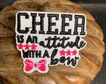 May include: A white patch with black embroidered text that reads "CHEER IS AN ATTITUDE WITH A BOW." The patch features pink stars and a pink bow design. The patch is on a wooden surface.