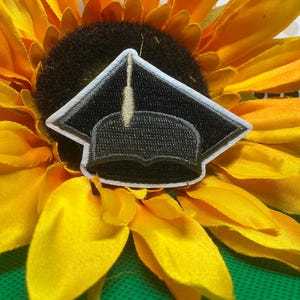 May include: A black embroidered graduation cap patch with a white border and a gold tassel detail. The patch features a square mortarboard top and a rounded cap base, showcasing detailed stitching throughout the design on a dark background.