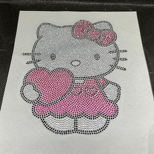 May include: A rhinestone-studded image of Hello Kitty holding a heart. The design features a pink bow, dress, and heart, with silver and black accents. The image is on a white background.