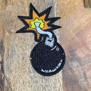 May include: A black and white embroidered patch of a cartoon bomb with a lit fuse. The bomb is round and has a white fuse with a yellow and orange flame.