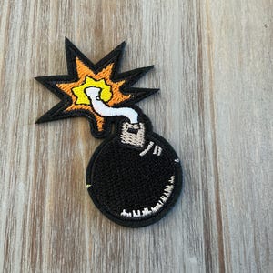 May include: Embroidered patch featuring a cartoon bomb with a black body and a white fuse. The explosion is depicted with yellow and orange flames, outlined in black. The patch is on a wooden surface.