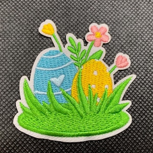 May include: Embroidered iron-on patch featuring two Easter eggs, one blue and one yellow, surrounded by green grass and pink and yellow flowers.