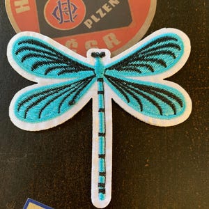 May include: Embroidered dragonfly patch with turquoise wings and black detailing. The patch has a white border and a black and turquoise body. The dragonfly is set against a dark background, with a vintage-style label visible.