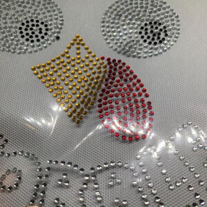 May include: A white fabric with a design of rhinestones. The design includes two silver circles with black centers, a gold diamond shape, and a red teardrop shape. The word "BEAUTY" is spelled out in silver rhinestones.