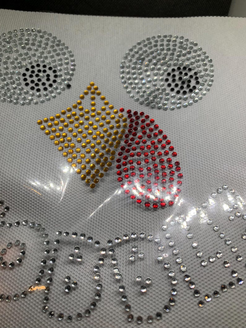 May include: A white fabric with a design of rhinestones. The design includes two silver circles with black centers, a gold diamond shape, and a red teardrop shape. The word "BEAUTY" is spelled out in silver rhinestones.