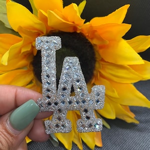 May include: A silver-colored "LA" patch with mirrored rhinestones. The patch is surrounded by yellow sunflower petals.
