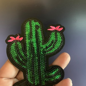 May include: A green cactus with pink flowers, made of sequins, is a decorative patch for clothing or crafts.