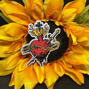 May include: A patch featuring a red heart pierced by two swords, with a crown of thorns and flames, set against a yellow sunflower. The patch has a white border and a black background.