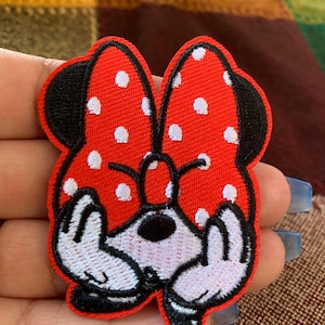 May include: A red and white polka dot Minnie Mouse bow-shaped patch with black trim. The patch features white gloved hands and a black nose. The patch is designed for application on clothing or accessories.