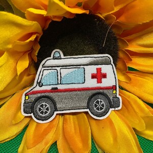 May include: Embroidered patch of an ambulance with a red cross on the side. The ambulance is gray, white, and red, with blue windows and black wheels. The patch is set against a bright yellow sunflower.