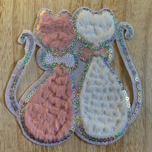May include: A decorative applique featuring two stylized cat figures. One cat is pink and fluffy, the other is white with a quilted texture. Both have sequined accents and bow ties, with a light gray outline.