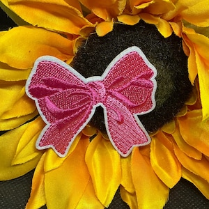 May include: A bright pink embroidered bow patch with a white outline, set against a vibrant yellow sunflower. The bow has a textured appearance, and the sunflower petals are a mix of yellow and orange.
