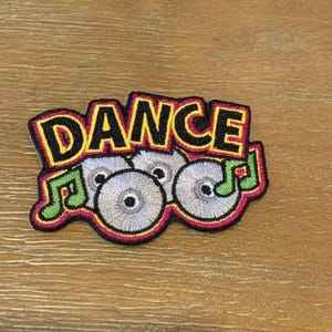 May include: A colorful embroidered patch with the word "DANCE" in black letters with yellow outlines. Three stylized silver record discs are below the word with green musical notes on either side.