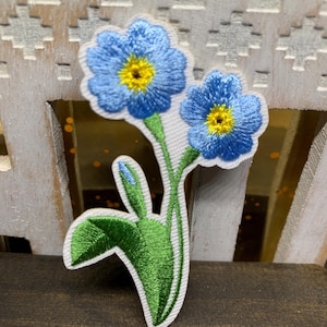 May include: Embroidered patch featuring two blue forget-me-not flowers with yellow centers and green stems and leaves. The patch has a white border and is set against a wooden background. The flowers are detailed with blue thread.