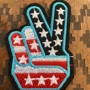 May include: A peace sign patch with a red, white, and blue American flag design. The patch is embroidered with white stars on a black background and has a blue outline.