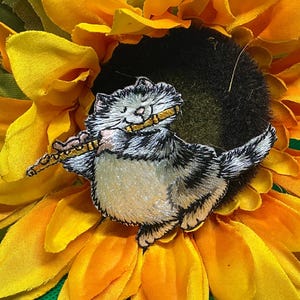 May include: Embroidered patch of a gray and white cat playing a flute, set against a bright yellow sunflower. The cat is detailed with black and white stripes and holds a golden flute. The sunflower has vibrant yellow petals and a dark center.