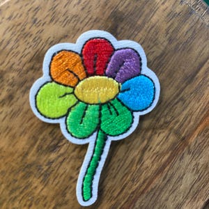 May include: Embroidered flower patch with a white border. The flower features petals in various colors: red, orange, yellow, green, blue, and purple. The stem is green. The patch is on a wooden surface.