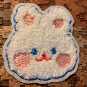 May include: A white fluffy bunny face patch with blue stitching around the edges. The bunny has pink cheeks, blue eyes, and a red nose.