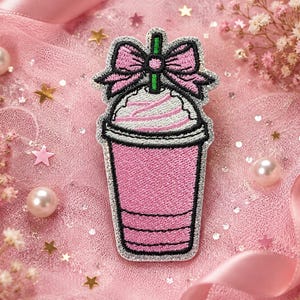 May include: An embroidered patch shaped like a pink beverage cup with a silver glitter border. The design features a pink cup, white whipped topping, a green straw, and a pink bow at the top, all outlined in black thread with detailed stitching.