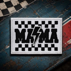 May include: A rectangular embroidered patch featuring a white background with black checkered borders at the top and bottom. The word MAMA is stitched in bold black letters, with a white lightning bolt replacing the letter A in the center.