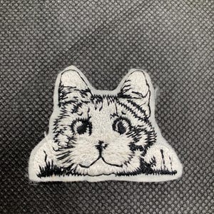 May include: Embroidered patch featuring a cat's face in white with black detailing. The cat's head is framed by its ears and paws, giving it a three-dimensional appearance. The patch is designed to be sewn or ironed onto fabric.